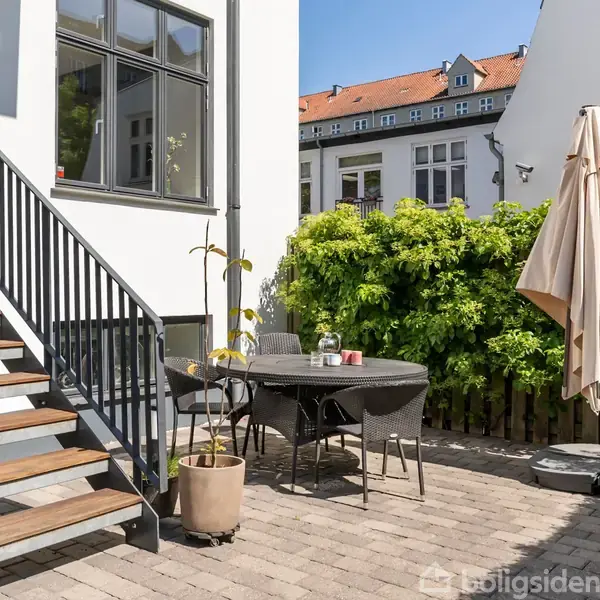 En udendørs terrasse med et rundt bord og stole, beliggende ved siden af en trappe. En stor parasol står til højre, med grønne buske og bygninger i baggrunden.