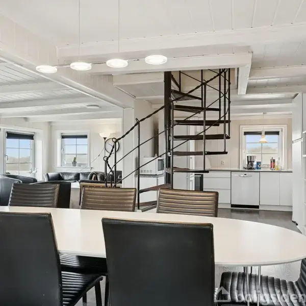 Rundt spisebord med sorte stole står i et lyst rum. En moderne spiraltrappe leder opad. I baggrunden ses en åben køkkenafdeling med hvide skabe og et opholdsområde med sofaer.