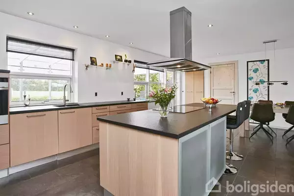 Køkken med lyse træskabe og en stor, sort køkkenø med en vase med blomster og en skål med frugt. Spisebord og stole ses i baggrunden ved vinduerne.