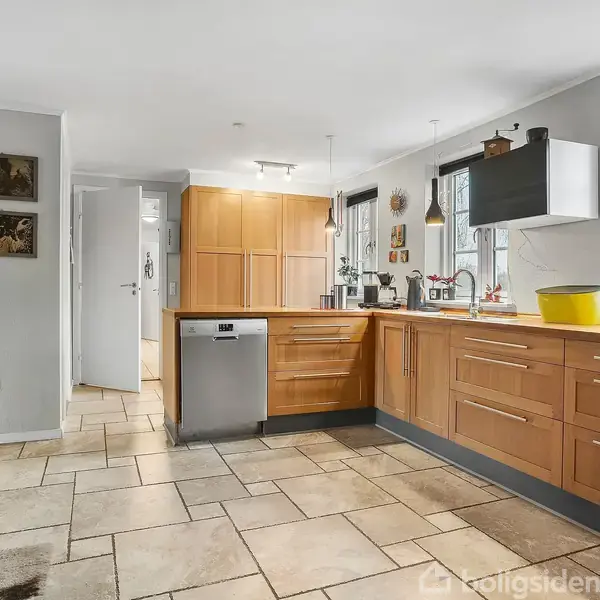 Et moderne køkken med træskabe og en opvaskemaskine. Køkkenet har lyse fliser og et stort vindue, der slipper lys ind. Dekorationer hænger på væggen, og der er køkkenredskaber fremme.