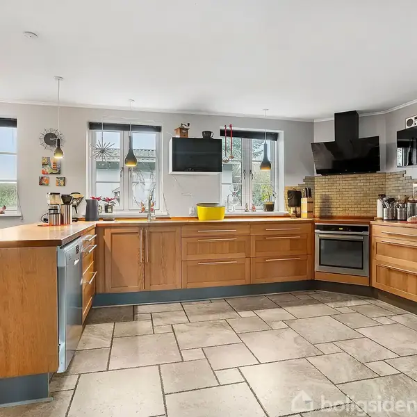 Et køkken med træskabe omkranser et værelse med tre vinduer. Der er en ovn, emhætte, opvaskemaskine og køkkenredskaber på bordpladerne. Stengulv og lysindfald gennem vinduerne.