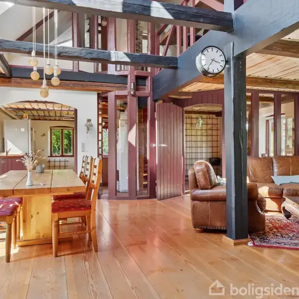 Et træspisebord står i en stue med højt til loftet, omgivet af træbjælker. Et ur hænger på en sort bjælke. Sofa og tæppe ses til højre.