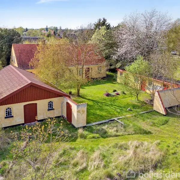 Gul bygning med rødt tag står stille omgivet af grønne træer og græs. En lille have med buske og en hvid blomstrende træ findes i forgrunden.