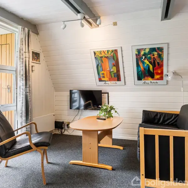 Stue med sort lænestol og sofa omkring et træbord med plante. Fladskærm hængende på væggen. Væggene er hvide med farverige billeder af "Aasy Madsen." Glasskydedør til terrasse.
