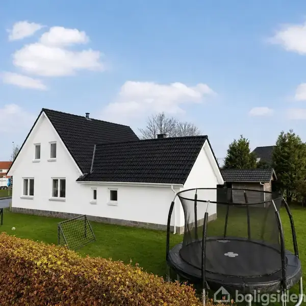 Hvidt hus med sort tag står i en have. En trampolin er placeret på græsplænen. Omgivelserne inkluderer hække, andre huse og en klar blå himmel med skyer.