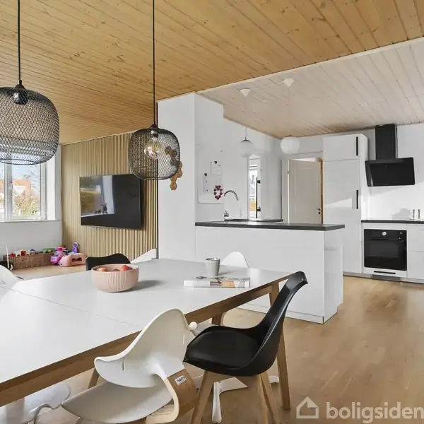 Spisebord med moderne stole og lamper i en lys stue-køkken alrum. Køkkenet er åbent med hvide skabe og sorte detaljer. Legetøj ses i baggrunden ved vinduet.