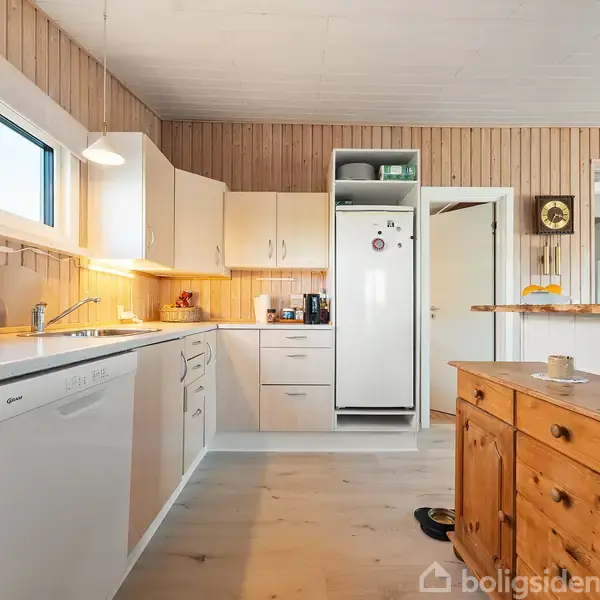 Køkken med hvidt køleskab og ovn, placeret ved en trævæg. Træskabe og lyse trægulve supplerer indretningen. Store vinduer og lamper giver lys til rummet, der er hyggeligt og funktionelt.