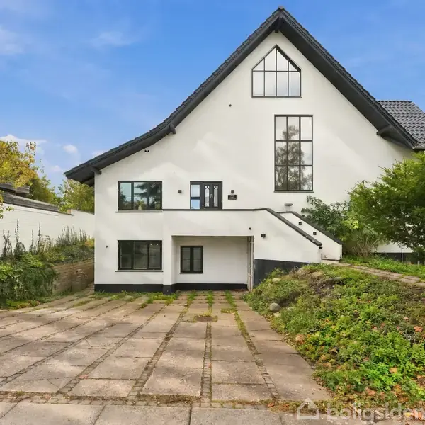 Hvid, moderne hus med sort tag ligger på en brolagt indkørsel. Trapper fører til hoveddør på første etage. Omgivet af grønne træer og buske, klar blå himmel.
