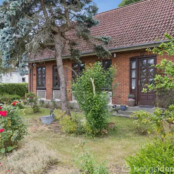 Et rødt murstenshus med en skråt tag og to indgangsdøre, omgivet af en velplejet have med blomster og træer. Græsplænen er grøn, og der er rosenbuske i forgrunden.