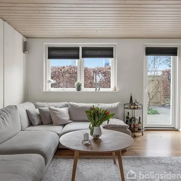 En grå hjørnesofa står i en stue ved et rundt sofabord med en vase med blomster. Rummet har lyst trægulv, træloft og store vinduer, der viser en have.