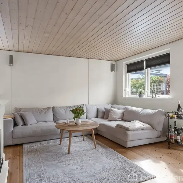 Grå hjørnesofa står i en lys stue med træloft, omgivet af hvide vægge og store vinduer. Foran sofaen står et rundt sofabord med blomster, mens en barvogn er ved siden af.
