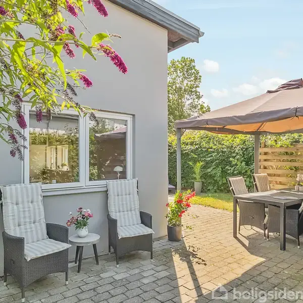 To stole med puder står foran et hus med store vinduer. Et havebord med parasol er placeret på en flisebelagt terrasse omgivet af grøn beplantning.