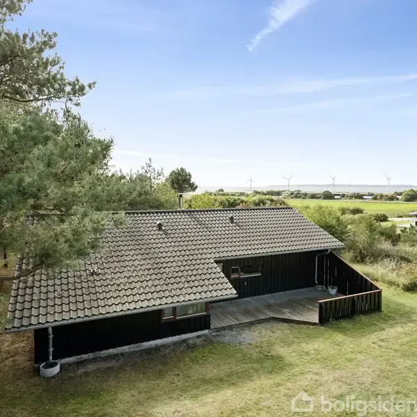 Sort træhus står stille omgivet af græs og træer, beliggende i et åbent landskab med udsigt til vindmøller i det fjerne under en blå himmel.