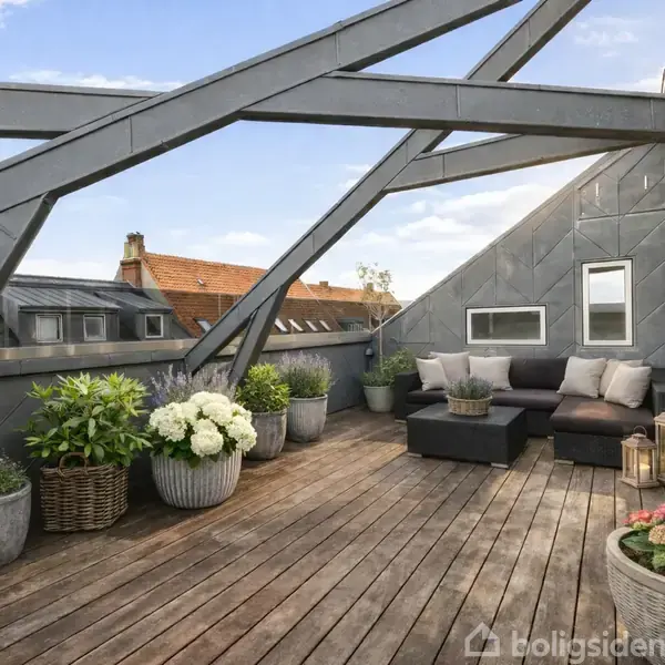 Terrasse med trægulv, hvor blomster i krukker står opstillet. En sofa med puder står langs væggen, omgivet af lanterner. Overdækning med metalkonstruktioner skaber et moderne udseende.