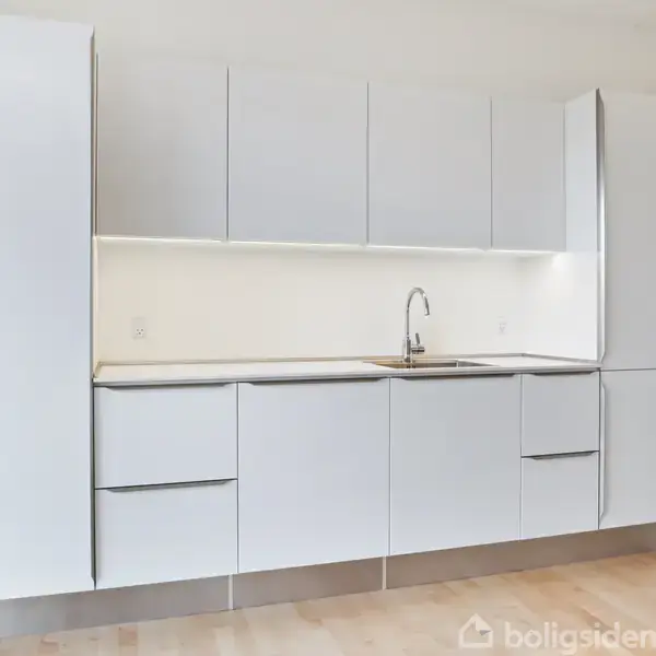 Et moderne, minimalistisk køkken med hvide skabe og skuffer. En vask er centralt placeret. Køkkenet er indbygget i en lys væg med et trægulv som underlag.