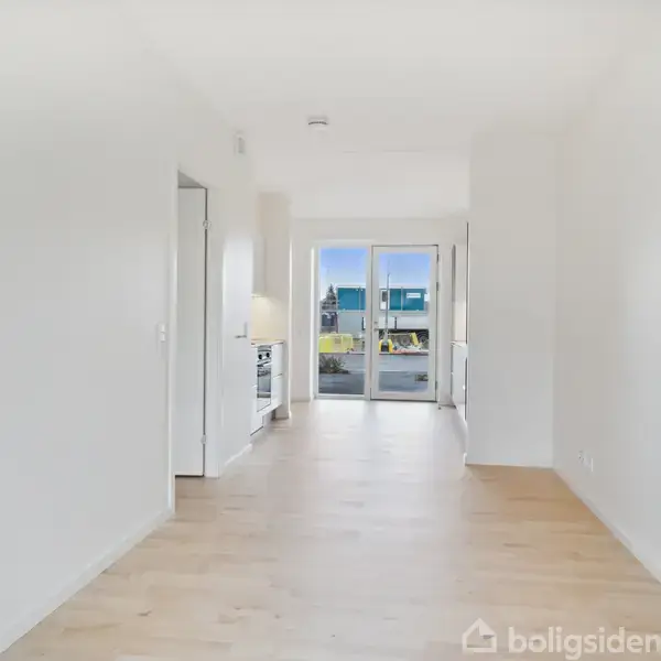 Langt, lyst værelse med trægulv fører til glasdøre, som åbner mod en balkon. Indgangen til et køkken ses til venstre. Store vinduer giver udsigt til området udenfor.