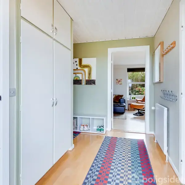 En entre med hvide skabe og trægulv har et farverigt gulvtæppe. Der er en åben dør til stuen foran, hvor man kan se sofaer og et stort vindue.