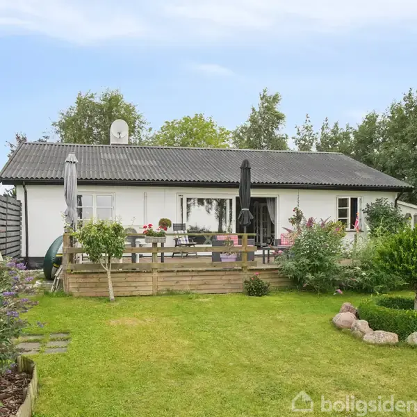 Et hvidt hus med sorte tagsten står omgivet af grønne træer og en velplejet græsplæne. Foran huset er en terrasse med stole, bord og planter.