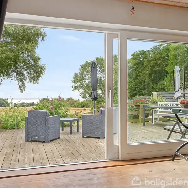 En træterrasse med havemøbler står stille, omgivet af grønne træer og blomster. Store glasdøre åbner til udendørsområdet, hvor landskabet fortsætter med udsigt til natur.