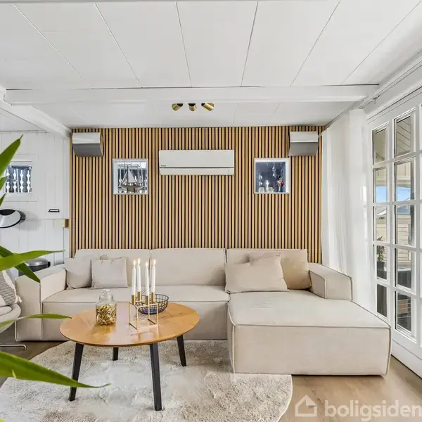 Stue med hvid sofa ved en væg med træpaneler, flankeret af to billeder og aircondition. Rundt træbord med lysestager i midten. Glasdøre åbner til udendørs terrasse. Plante i forgrunden.