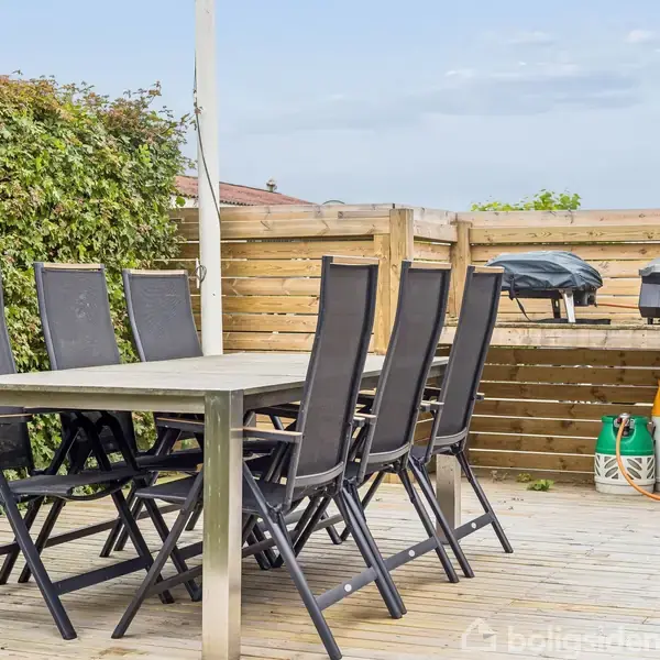 Et havebord med seks sorte stole står på en træterrasse. En grill er placeret på en hylde ved et træhegn. Grønne planter omkranser området.
