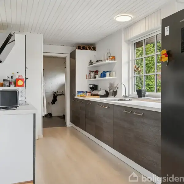 Et moderne køkken med hvide vægge og skabslåger i mørkt træ. Et køleskab står til højre, mens en mikrobølgeovn og blender er placeret på køkkenbordet. Vinduet er på højre væg.