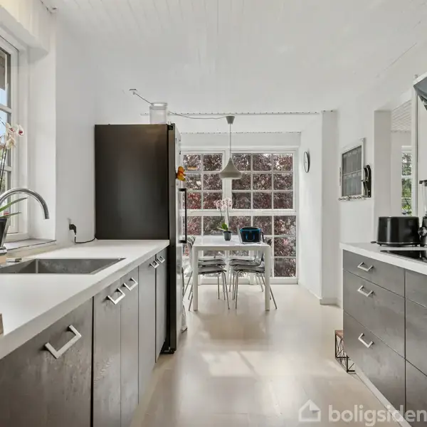 Et moderne køkken med mørke skabe flankerer en lys spiseplads ved vinduet. Udenfor ses grønne træer. En skærm og kaffemaskine er til stede på bordet.