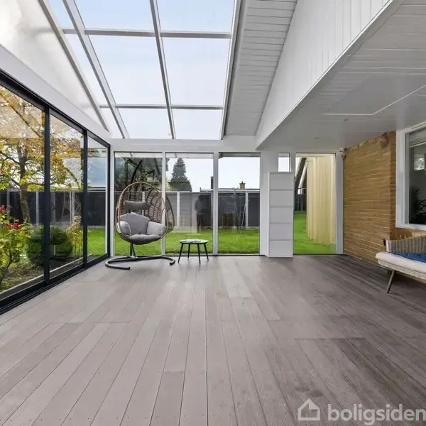 Værelse med glasvægge og træloft har en hængestol og et bord. Vinduet viser udsigt til en grøn have med træer og blomster. Skandinavisk minimalistisk stil.