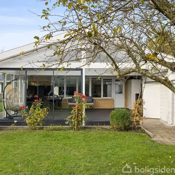 Hus med glasudestue, omgivet af grøn, velplejet have. En hængestol ses i udestuen. Træer og buske i haven skaber en fredelig atmosfære.