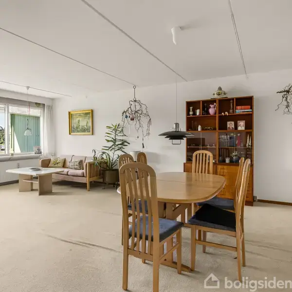 Spisebord med seks stole står i en stue, hvor der i baggrunden er en sofa og et bogreol. Store vinduer med udsigt til en grøn have.