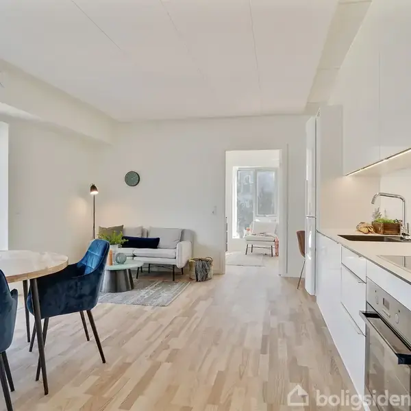 Et lyst køkken-alrum har et rundt spisebord med blå stole, en sofa og et åbent køkken. Rummet er stilfuldt møbleret med lyse trægulve og moderne inventar.