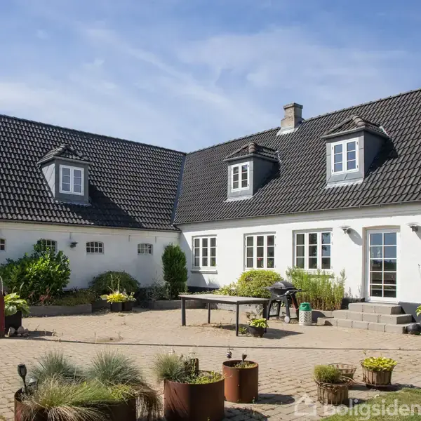 Et hvidt hus med sort tag med kviste står foran en flisebelagt gårdhave; der er planteliv i potter og en græsplæne i baggrunden under en klar blå himmel.