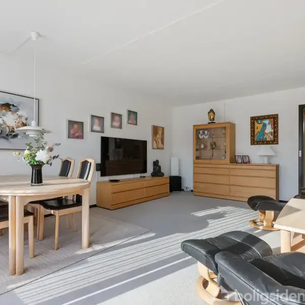 Stue med spisebord og fire stole til venstre, TV stående på lavt møbel, og reol med skuffer til højre. Omgivet af kunstværker på væggene.