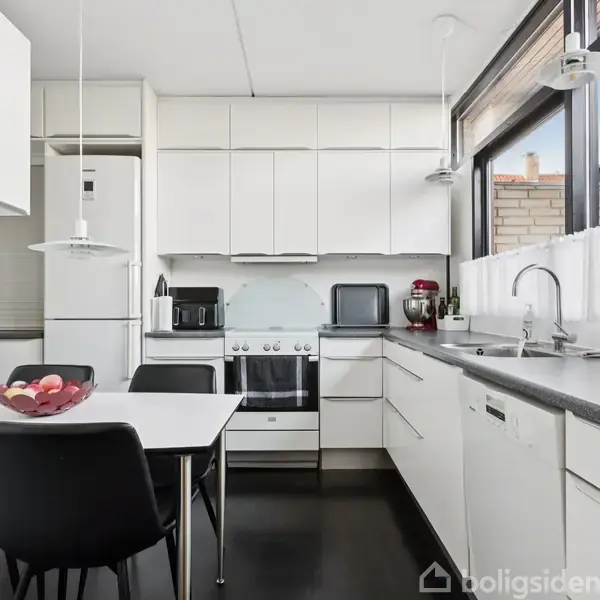 Køkkenbord står i midten af et moderne køkken med hvidt interiør. En skål frugt står på bordet. Store vinduer til højre giver naturligt lys, omgivet af hvide skabe og apparater.