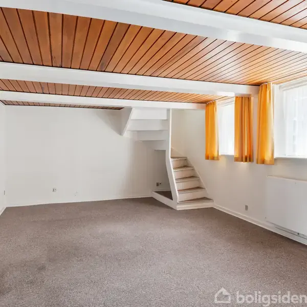 Et rum med grå tæppebelægning, orange gardiner i vinduerne og træloft. En hvid trappe fører opad. Væggene er hvide, og der er en radiator under vinduerne.