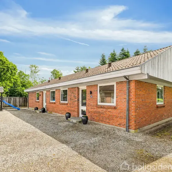 Et rødt murstenshus med hvide vinduer står på en gårdsplads med grus. Omgivelserne inkluderer grønne træer og en legeplads med en rutsjebane til venstre.