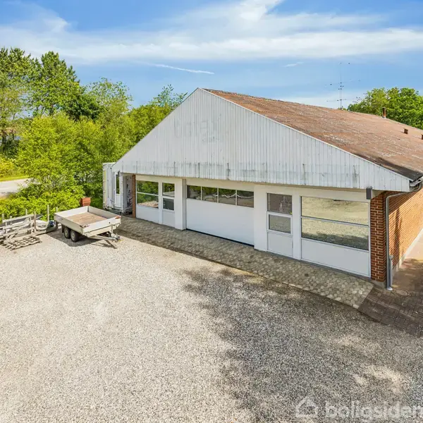 Bygning med hvid facade og store vinduer står stille i grusbelagt område. Den omkringliggende kontekst er frodig med grønne træer og en klar blå himmel.