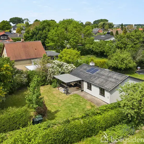 Hus med solpaneler på taget står i en frodig have, omgivet af grønne træer og buske. Andre huse med røde tage ses i nabolaget under en klar himmel.