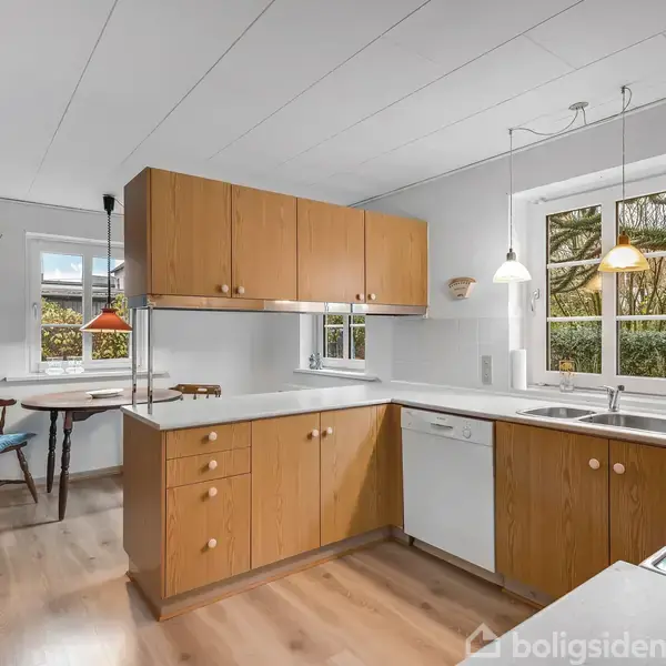 Køkken i lyst træ med hvide overflader. Skabe hænger over en opvaskemaskine ved et vindue. En lille spiseplads med to stole står ved et andet vindue. Lamper hænger fra loftet.