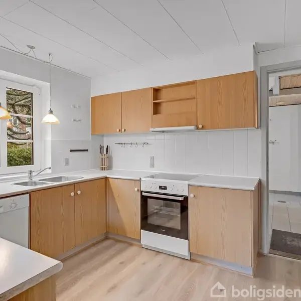 Et lyst køkken med træskabe og hvide bordplader. Komfuret står ved en væg, mens en vask og opvaskemaskine er under vinduet, der har udsigt til en grøn have.