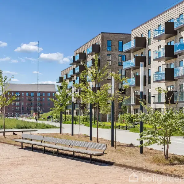 Flere etagebygninger med altaner står langs en flisebelagt sti. Foran bygningerne er der bænke, træer og cykelstativer, alt under en klar blå himmel.