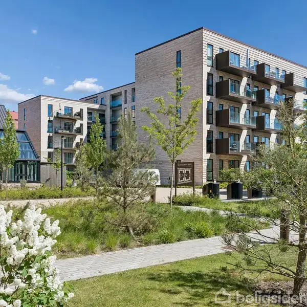 Bygning står stille, omkranset af grønne træer og blomster, med altaner langs facaden. Baggrund har en blå himmel med spredte skyer, skaber en fredelig atmosfære.