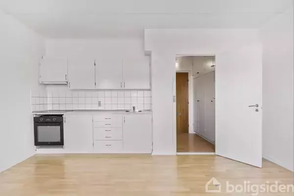 Et køkken i en lejlighed med hvide skabe og skuffer, en indbygget ovn, og vask placeret langs en hvid væg med fliser. Åben dør fører til et andet rum.