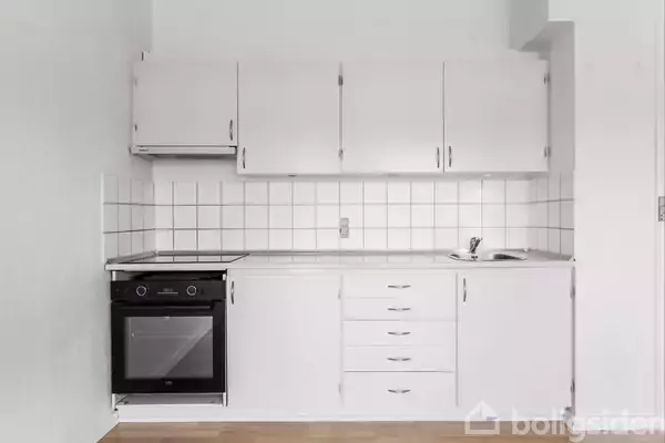 Et hvidt køkken med ovn og komfur integreret i skabene. Flisevæggen er bagved, og en lille vask er til højre på bordpladen. Gulvet er trægulv.