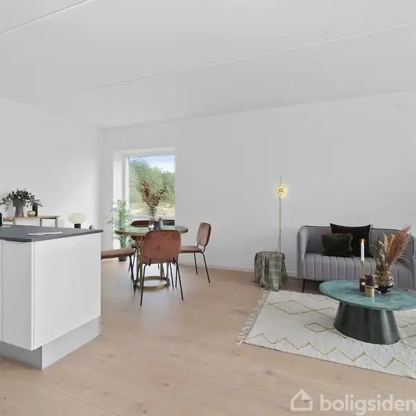 Et moderne køkken-alrum med hvide elementer og en sort bordplade. Til venstre står spisebord med stole, til højre en grå sofa ved side af et grønt sofabord. Rummet er lyst med store vinduer.