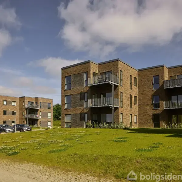Flere moderne lejlighedskomplekser står på en græsplæne under en blå, let skyet himmel. Bygningerne er i brun mursten med glasaltaner, og nogle biler er parkeret i baggrunden.