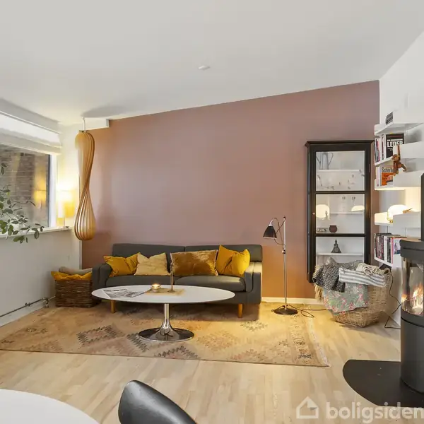 Stue med sofa og sofabord foran en varm, brun væg. Pejs til højre ved siden af en reol. Vinduer til venstre slipper naturligt lys ind. Gulvtæppe og planter tilføjer hygge.