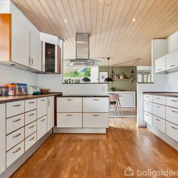 Køkken med hvide skabe og skuffer, to ovne indbygget i væggen, træloft og trægulv. Der er lys gennem vinduet, og spiseområdet ses i baggrunden.