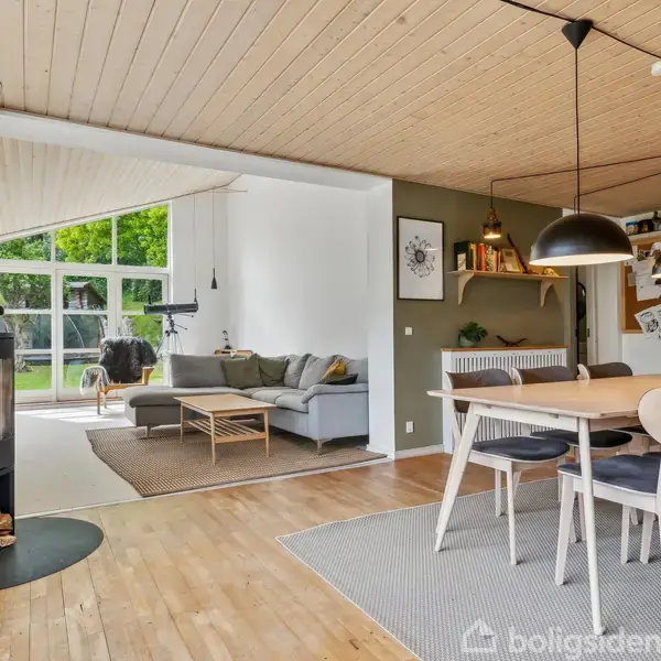 Brændeovn står tændt i en stue med trægulv. Ved siden af er et gråt sofaområde med stort vinduesparti. Spisebordet er dækket under to sorte lamper.