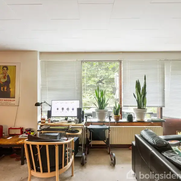 En stue med sofaer og en computerstation ved et stort vindue, omgivet af potteplanter. Væggen har en plakat med teksten "Henry Heerup". En rød lampe hænger ved siden af røde sofaer.